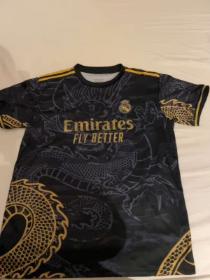 Real Madrid Ronaldo tröja svart/guld - Snygg Real Madrid matchtröja från Adidas i svart med guldiga detaljer och drakmönster. Tröjan har gula ränder på axlarna, klubbmärke på bröstet och texten 'Emirates Fly Better' framtill. På ryggen står det 'Ronaldo 7' i gult.
