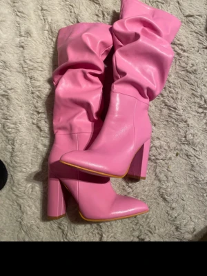 Rosa höga stövlar med klack - Säljer ett par höga stövlar i stark rosa färg med bred klack och spetsig tå. Stövlarna har en slouchy design och dragkedja på insidan. Tillverkade i ett glansigt syntetmaterial som ger en trendig look.