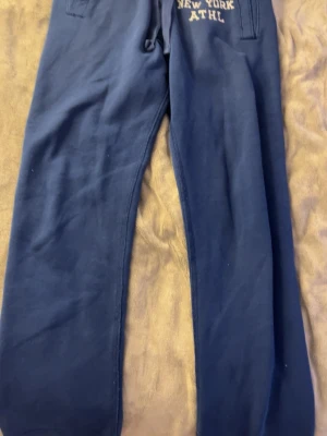 Mörkblå mjukisbyxor från A&F - Mörkblå mjukisbyxor från Abercrombie & Fitch med broderad text 'A&F New York ATHL' i gult vid fickan. Byxorna har dragsko i midjan och två sidofickor. Perfekta för chill dagar och riktigt sköna att ha på sig.