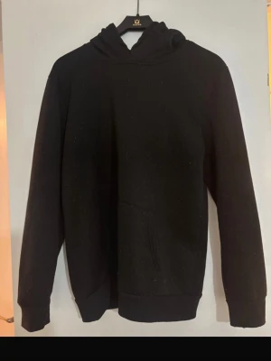 Svart hoodie med huva och ficka - En klassisk svart hoodie med huva och dragsnören, tryck på ryggen. Hoodien har en stor magficka framtill och ribbade muddar vid ärmslut och nederkant. Perfekt för en avslappnad och stilren look. Materialet känns mjukt och bekvämt, troligen bomullsblandning.