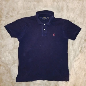 Mörkblå pikétröja från Polo Ralph Lauren - Klassisk mörkblå pikétröja från Polo Ralph Lauren med korta ärmar och krage. Tröjan har knappar framtill och den ikoniska röda logotypen broderad på bröstet. Perfekt till jeans eller shorts för en clean och stilren look. Har en väldigt svag vit fläck längst ner typ på piken