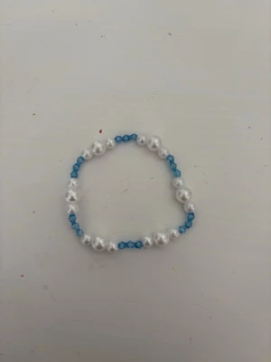 Pärlarmband med blå detaljer - Snyggt armband med vita pärlor och små blå glaspärlor i ett enkelt, elastiskt band. Perfekt för att addera lite färg till din outfit. Armbandet har en klassisk rund form och är handgjort med pärlor i plast.