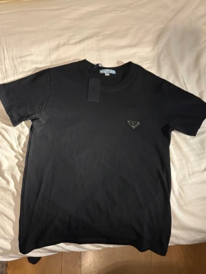 Svart t-shirt från Prada - Säljer en svart t-shirt från Prada med klassisk rund hals och korta ärmar. T-shirten har en diskret Prada-logga i form av en metalltriangel på bröstet. Materialet är mjuk bomull och passformen är normal. Perfekt för dig som gillar stilrena designer. Lappen sitter kvar är i M men passar s ochså