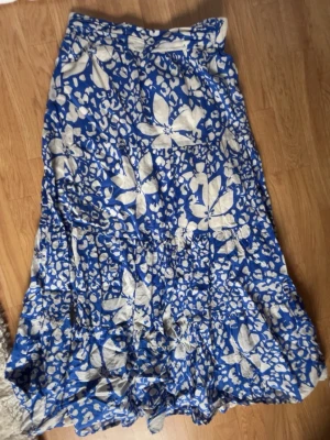 Blå långkjol med blommigt mönster💗 - Snygg blå långkjol med vitt blommigt mönster och volangdetaljer. Kjolen har en luftig och flowy passform, perfekt för varmare dagar. Den har en elastisk midja och är tillverkad i ett lätt material som känns bekvämt hela dagen.