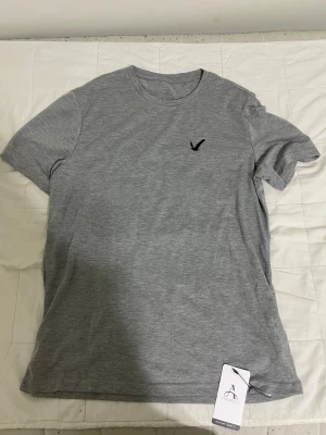 Grå t-shirt från OVS med broderad logga - Snygg grå t-shirt från OVS i mjuk bomull. T-shirten har en klassisk passform, rund halsringning och korta ärmar. På bröstet finns en diskret svart broderad logga i form av en fågel. Perfekt basplagg till garderoben.
