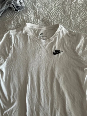 Vit Nike T-shirt med logga - Klassisk vit T-shirt från Nike med svart broderad logga på bröstet. Tillverkad i mjuk bomull och har rund halsringning samt korta ärmar. Perfekt basplagg för en clean och sportig stil.