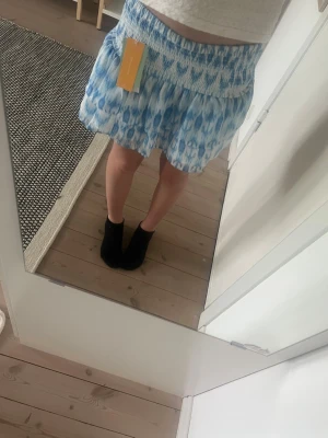 Blåvit kortkjol med volang - Supersöt kortkjol i blått och vitt med volang och smock i midjan. Kjolen har ett mönster som påminner om batik och är perfekt för varma dagar. Den har en luftig och flowy passform som ger en avslappnad vibe. Storlek 146/152 från Gina 