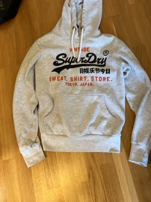 Ljusgrå Superdry hoodie med tryck - Snygg ljusgrå hoodie från Superdry med stor logga och text i svart, rött och vitt på bröstet. Klassisk känguruficka framtill, dragsko i huvan och mjukt material. Perfekt för en avslappnad streetstyle-look.