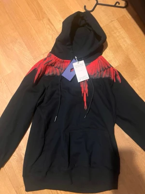 Svart hoodie Marcelo Burlon med röda vingar - Passar M-L .Svart hoodie från Marcelo Burlon County of Milan med ikoniskt vingtryck i rött och grått över axlar och bröst. Klassisk huva med dragsko och stor magficka framtill. Perfekt statement-plagg för dig som vill sticka ut.