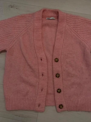 Rosa kofta från Zara med knappar - Mysig rosa kofta från Zara med grovstickad struktur och fyra bruna knappar framtill. Koftan har ribbade muddar och en djup v-ringning. Perfekt att slänga på sig över en t-shirt eller linne.