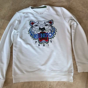 Vit Kenzo sweatshirt med tigertryck - Vit sweatshirt från Kenzo med ikoniskt broderat tigerhuvud i blått, rött och svart framtill. Rund halsringning och ribbade muddar. Perfekt för dig som gillar streetwear och vill sticka ut med en klassisk design.