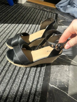 Svarta kilklackade espadrillesandaler - Säljer ett par svarta sandaler med kilklack och breda remmar över foten. Sandalerna har justerbart ankelband med guldfärgat spänne och klacken är täckt med flätad jute för en klassisk espadrille-look. Perfekta för sommaren och ger extra höjd utan att tumma på komforten.