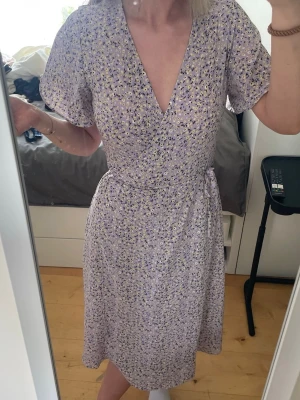 Ljuslila blommig omlottklänning - Supersöt ljuslila omlottklänning med småblommigt mönster i vitt och gult. Klänningen har korta ärmar, v-ringning och knyts i midjan för en smickrande passform. Perfekt för vår och sommar!🥰
