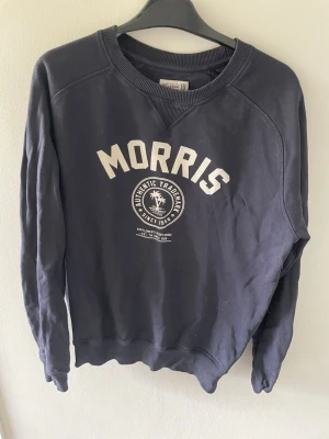Svart sweatshirt från Morris - Klassisk svart sweatshirt från Morris med tryckt logga och text på bröstet. Tröjan har rund halsringning, ribbade muddar vid ärmslut och nederkant samt en avslappnad passform. Perfekt för en casual look.