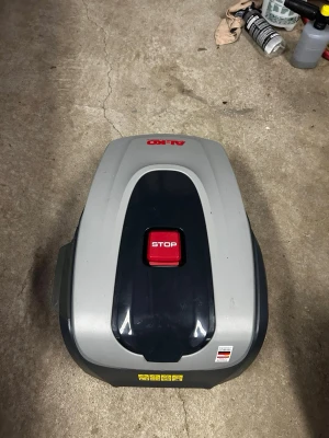 AL-KO Robolinho 500 E robotgräsklippare - Robotgräsklippare från AL-KO, modell Robolinho 500 E. Färgen är grå med svarta och röda detaljer, och den har en tydlig röd STOP-knapp på ovansidan. Maskinen har en modern, rundad design och är tillverkad i tålig plast. Perfekt för automatiserad gräsklippning.