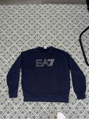 Mörkblå EA7 Emporio Armani sweatshirt - Snygg mörkblå sweatshirt från EA7 Emporio Armani med stort broderat logotyptryck på bröstet. Tröjan har rund halsringning, långa ärmar och ribbade muddar. Perfekt för en stilren och avslappnad look.
