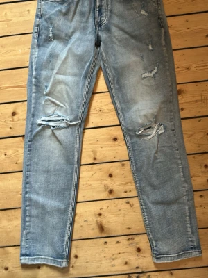 Ljusblå slitna jeans med raka ben - Säljer ett par ljusblå jeans med slim ben och flera slitningar framtill för en trendig, avslappnad look. Jeansen har klassisk femficksdesign och är tillverkade i denim. Perfekta för dig som gillar streetstyle och vill ha en cool vibe. Pris kan diskuteras och de är bara att fråga på!