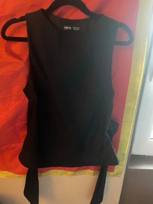 Svart knyt topp från SHEIN - Stilren svart ärmlös topp från SHEIN med breda axelband och snygg knytning i midjan. Toppen har en rak passform och är perfekt för att skapa en clean och trendig look. Materialet känns mjukt och lätt, troligen polyester.