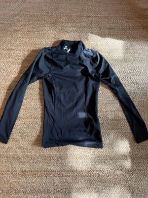 Träningströja Under Armour - Marinblå långärmad träningströja från Under Armour med hög krage och logga på halsen. Tillverkad i ett stretchigt funktionsmaterial som andas (musclefit). Knappt använd.