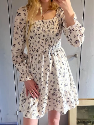 Vit blommig kortklänning med smock - Supersöt vit kortklänning med blommönster och smockad överdel. Klänningen har långa ärmar med lätt volang vid ärmslut och en fyrkantig ringning. Perfekt för vår och sommar.