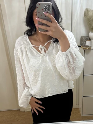 Vit blommig blus med knytband - Gina Tricot L - Söt vit blus med blommig struktur och transparenta långärmar. Blusen har ett knytband vid halsen och avslappnad passform. Perfekt för dig som gillar romantisk stil. Materialet känns lätt och luftigt. Köptes från Gina Tricot, storlek L. Helt ny, oanvänd.