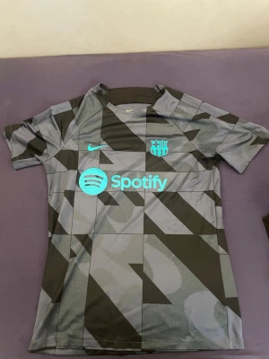 Barcelona matchtröja Nike grå/svart - Snygg Barcelona matchtröja från Nike i grått och svart med geometriskt mönster. Tryck med Spotify-logga och klubbmärke i turkos på bröstet. Korta ärmar och sportig passform, perfekt för fotboll eller träning.