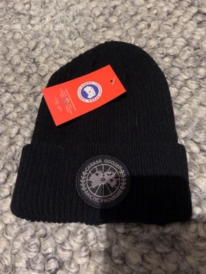 Svart ribbstickad mössa Canada Goose - Svart ribbstickad mössa från Canada Goose med uppvikt kant och rund patch framtill med logga och texten 'Arctic Program'. Klassisk och stilren modell som passar perfekt till kalla dagar.
