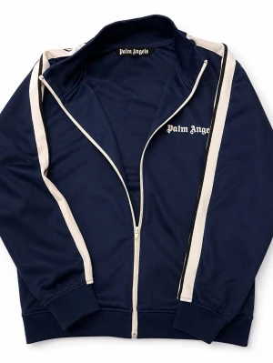 Palm Angels marinblå zip-tröja - Palm Angels marinblå zip-tröja med vita kontrastpaneler längs ärmarna och vit dragkedja. Klassisk hög krage och logga på bröstet. Tillverkad i ett mjukt, sportigt material som ger en avslappnad streetwear-look.