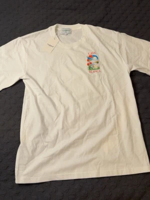 Vit Casa Blanca t-shirt  - Vit t-shirt från Casablanca med färgglatt tryck av rosor och landskap på ryggen samt mindre tryck på bröstet. Klassisk passform och rund halsringning. Perfekt för dig som gillar stilrena plagg med unik design. STORLEK L, PASSAR L OCH XL. HELT NY, OANVÄND.