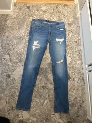 Blå Jeans - Säljer ett par ljusblå jeans från hollister med slitningar och hål på båda benen. Jeansen har klassisk femficksmodell, låg midja och är tillverkade i stretchigt denimtyg för en bekväm passform. Perfekta för en avslappnad och trendig look.