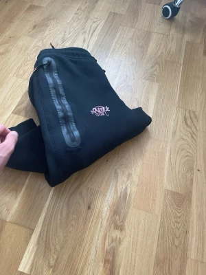 Svart Nike hoodie med rosa logga - Säljer en svart mjukis byxor från Nike med en snygg rosa Nike-logga på. . Materialet är mjukt och bekvämt, perfekt för att chilla eller hänga med kompisar. En cool detalj är den diskreta grafiska designen på framsidan.