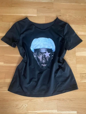 Svart t-shirt med Tyler the creator tryck - Svart t-shirt med rund hals och korta ärmar. Framtill finns det ett Tyler The Creator tryck som ger en cool kontrast till den svarta basen. T-shirten har en avslappnad passform och är tillverkad i mjukt material, perfekt för en casual look. Har klippt av halsringen. 
