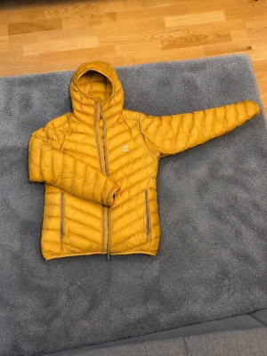 Gul dunjacka från Haglöfs med huva - Snygg gul dunjacka från Haglöfs med fast huva och två dragkedjeförsedda fickor framtill. Jackan har quiltad design och smidig passform, perfekt för kalla vinterdagar. Liten logga på bröstet och hel dragkedja framtill.