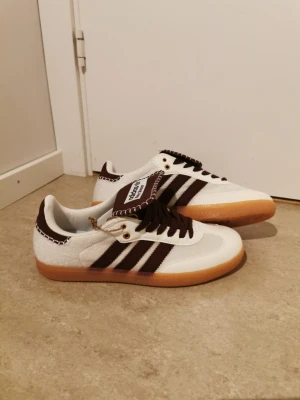 Adidas Wales Bonner vita sneakers - Unika Adidas Wales Bonner sneakers i vitt med bruna detaljer och klassiska tre ränder. Skorna har brun plös med vit kontrastsöm, bruna skosnören och gummisula. Materialmix av läder och textil ger en retrovibe och cool look.