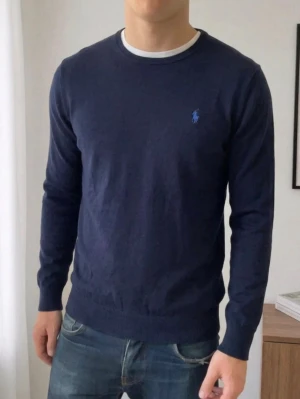 Mörkblå stickad tröja Polo Ralph Lauren - Mörkblå stickad tröja från Polo Ralph Lauren med rund halsringning och diskret logga broderad på bröstet. Tröjan har en klassisk passform och är perfekt att styla med jeans eller skjorta under.