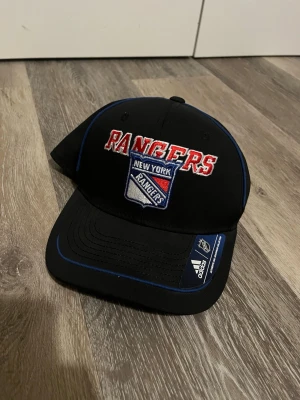 Svart New York Rangers keps Adidas - Snygg svart keps från Adidas med New York Rangers broderad logga och text i rött, vitt och blått. Böjd skärm med blå detaljer och officiell NHL-märkning. Perfekt för dig som gillar hockey och streetwear. Pris kan absolut diskuteras.   