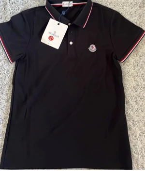 Svart polotopp från Moncler - Säljer en svart polotopp från Moncler med klassisk krage och korta ärmar. Toppens krage och ärmslut har snygga rödvita detaljer. Moncler-loggan sitter på bröstet. Perfekt för dig som gillar stilrena och sportiga plagg.