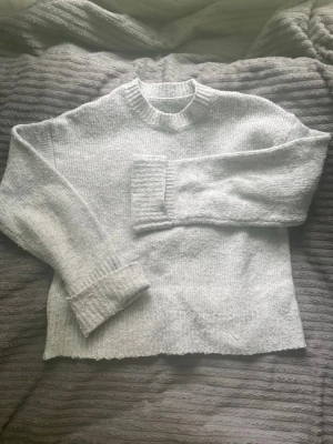 Ljusgrå stickad tröja med rund hals - Mysig ljusgrå stickad tröja med rund halsringning och ribbade muddar vid ärmslut och hals. Tröjan har en rak passform och är tillverkad i ett mjukt, melerat garn som ger en avslappnad look. Perfekt för lager-på-lager-stil under kyliga dagar.