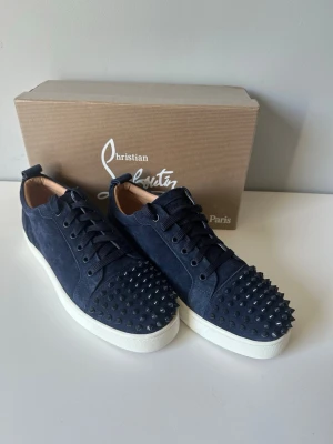 Mörkblå Christian Louboutin sneakers - Snygga mörkblå sneakers från Christian Louboutin i mocka med svarta nitar över tån. Skorna har vita sulor, svarta skosnören och en klassisk låg modell. Perfekta för dig som vill sticka ut med en edgy och lyxig stil.