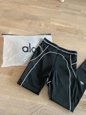 Svarta leggings från Alo med vita detaljer - Snygga svarta träningsleggings från Alo Yoga.. Kommer med originalpåse. Säljes pga de är för små i storleken. Prislapp finns kvar. 