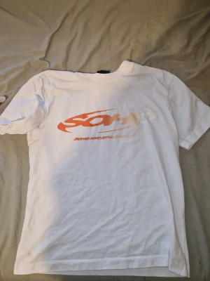 Vit t-shirt med orange Soho-tryck - Vit t-shirt från Soho NYC Athletic med ett stort orange och vitt tryck på bröstet. Klassisk passform med rund hals och korta ärmar. Perfekt för en avslappnad streetwear-look.