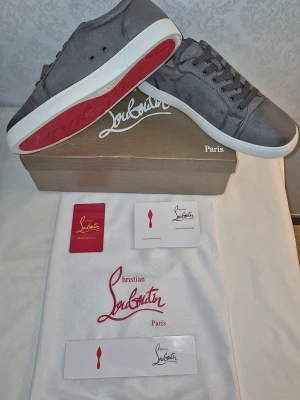 Gråa Christian Louboutin sneakers - Snygga gråa sneakers från Christian Louboutin med klassisk röd sula och vita kanter. Skorna har en stilren design i mocka med matchande gråa skosnören och diskreta sömmar. Perfekta för dig som vill ha en lyxig och trendig look.