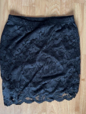 Svart spetskjol med blommigt mönster - Snygg svart kortkjol med blommigt spetsmönster och elastisk midja. Kjolen har en feminin och elegant look med vågig nederkant. Perfekt att styla med en enkel topp eller stickad tröja.