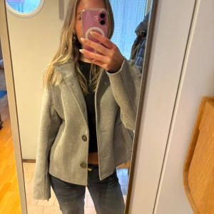 Grå kappa med knappar - Säljer en stilren grå kappa med knappar framtill. Jackan har en klassisk krage och långa ärmar, perfekt för kyligare dagar. Passar bra till både jeans och klänning.