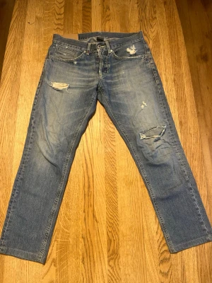 Blå dondups jeans - Ett par blå jeans med slitningar och hål på både fram- och baksida. Jeansen har klassisk femficksmodell, raka ben och en tvättad look. Perfekta för en avslappnad och trendig stil. Dondup!