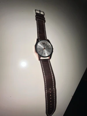 Emporio Armani brun klocka - Stilren klocka från Emporio Armani med silverfärgad urtavla och brunt läderarmband med krokodilpräglad struktur. Klockan har roséguldsdetaljer och klassisk design, perfekt för dig som gillar en tidlös accessoar. Min pappas gamla kan inte mycket om den batteriet funkar inte måste bytas ut  och banden kanske lite.