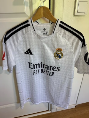 Real Madrid matchtröja Bellingham - Säljer en vit Real Madrid matchtröja från Adidas med svarta detaljer och klubbmärke på bröstet. Tröjan har Bellingham och nummer 5 på ryggen samt sponsortryck på framsidan. Tillverkad i lätt och ventilerande material, perfekt för fotbollsträning eller match.