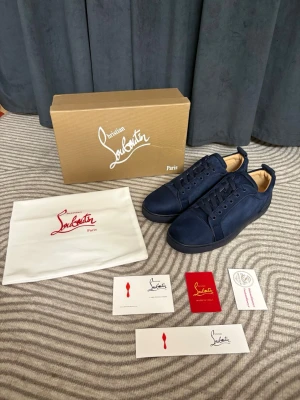 Marinblå sneakers från Louboutin - Snygga marinblå sneakers från Christian Louboutin i mocka med ton-i-ton broderad logga på hälen. Skorna har klassisk låg siluett, matchande snörning och svart sula. Perfekta för dig som vill ha en stilren och exklusiv look.