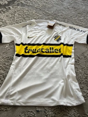 AIK vit matchtröja Nike - AIK:s vita matchtröja med svart och gul detalj över bröstet och Truecaller som sponsortryck. Tröjan har klubbmärke på vänster bröst, Nike-logga på höger och tryck på ärmarna. Tillverkad i lätt och ventilerande material, perfekt för fotboll eller träning.