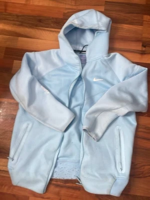 Ljusblå NOCTA Nike hoodie med dragkedja - Snygg ljusblå hoodie från Nike NOCTA-kollektionen. Hoodien har dragkedja framtill, två fickor med dragkedja och en klassisk huva. Tillverkad i mjukt material som känns skönt mot huden. Diskret Nike Swoosh på bröstet och NOCTA-logga på ärmen.
